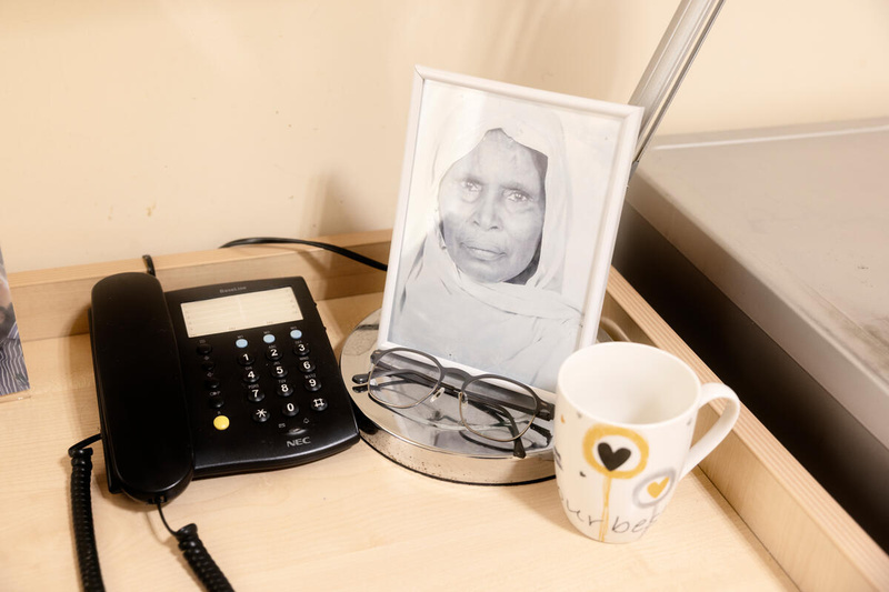 A framed black and white photo of an older woman sits on a desk next to a phone, amug and a pair of reading glasses