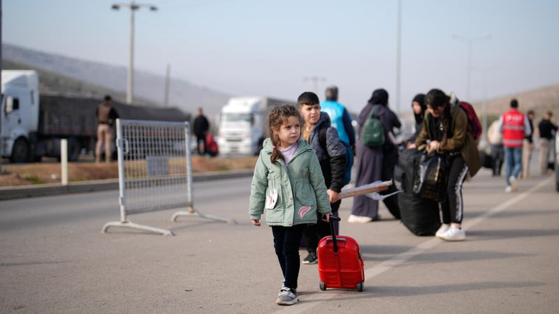 A young Syrian refugee child pulls a small suitcase along a road, heading towards a border crossing.