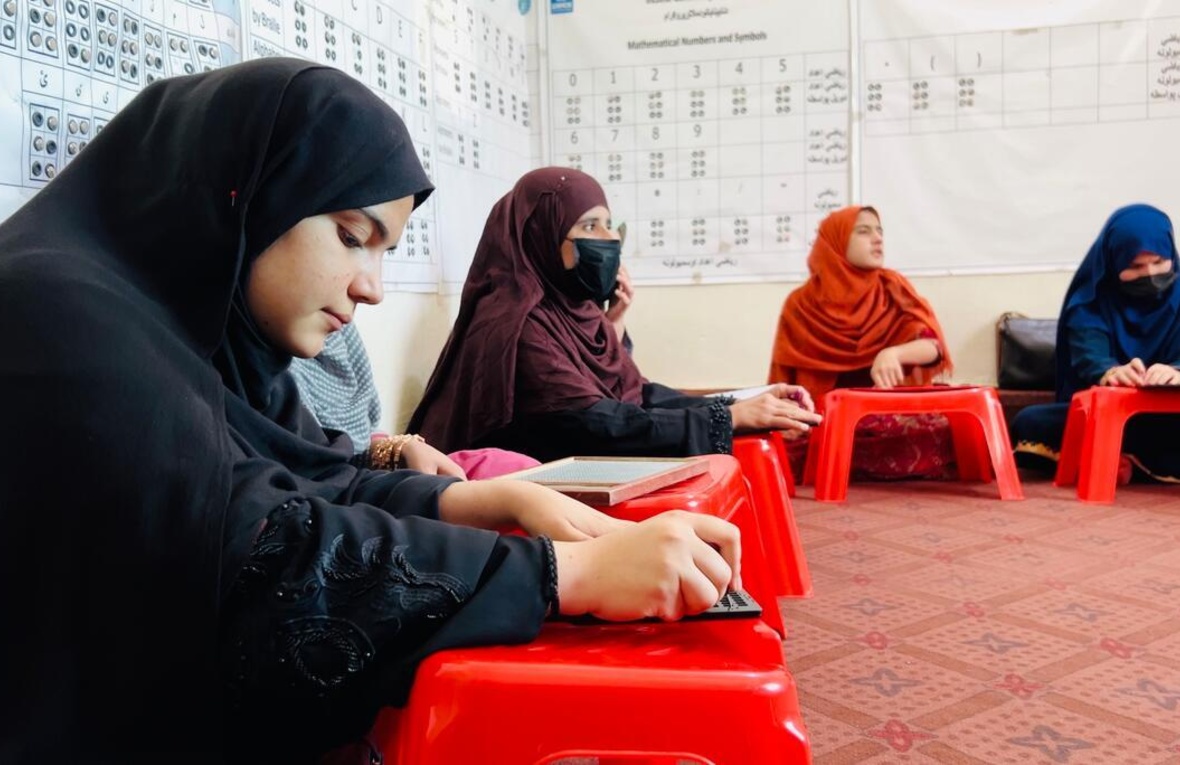 Helai Noori sits on the floor of a classroom with other women and reads Braille on a wooden tablet.