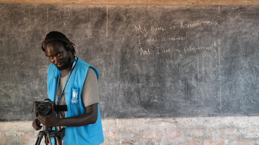 A man wearing a blue vest operates a camera in front of a blackboard with the writing: "I am Achouth. I was a refugee. And I am a filmmaker."