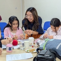 A facilitator stands beside a table, helping three children and a woman wearing a headscarf with a hands-on craft activity. The group is focused on working with yarn and small tools, while balls of pink and red yarn, cups, and activity materials are spread across the table. The workshop takes place in a bright room with blue chairs arranged along the wall.