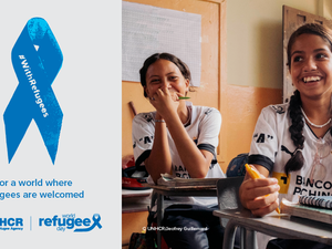 On the right, a picture of two young girls smiling and sitting at their desks in a classroom. On the left, a blue ribbon with the inscription "With Refugees". Text under the ribbon reads: "For a world where refugees are welcome".