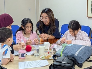 A facilitator stands beside a table, helping three children and a woman wearing a headscarf with a hands-on craft activity. The group is focused on working with yarn and small tools, while balls of pink and red yarn, cups, and activity materials are spread across the table. The workshop takes place in a bright room with blue chairs arranged along the wall.
