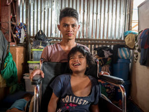 A young man kneels behind a boy who sits in a wheelchair. 