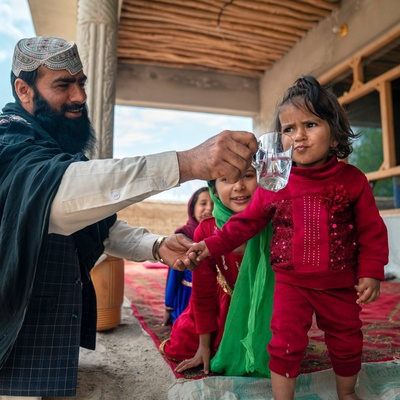 A man smiles as he holds a glass of water for a young girl to drink from.