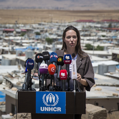 Angelina Jolie stands at a podium covered with news microphones, with Domiz refugee camp in the background