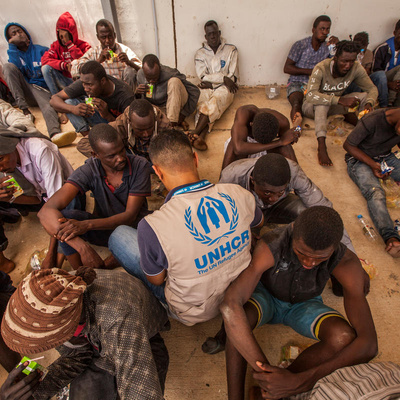 A group of disembarked refugees and migrants sit on the floor, joined by a UNHCR staff member wearing a tan UNHCR vest with blue logo.