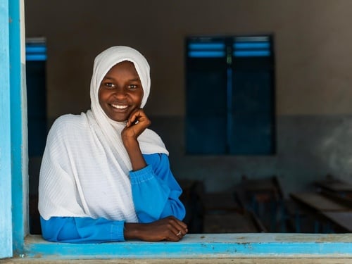 A young girl smiles at the camera, looking through an open window.