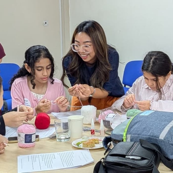 A facilitator stands beside a table, helping three children and a woman wearing a headscarf with a hands-on craft activity. The group is focused on working with yarn and small tools, while balls of pink and red yarn, cups, and activity materials are spread across the table. The workshop takes place in a bright room with blue chairs arranged along the wall.