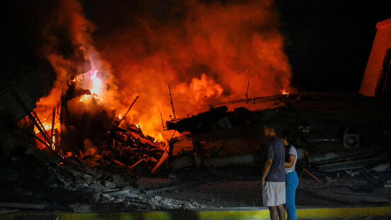 Two persons standing together at night, looking toward a large fire consuming the remains of damaged structures. Bright orange flames and thick smoke illuminate the scene, while debris is scattered across the ground. The image conveys the impact of an earthquake and the uncertainty faced by those affected.