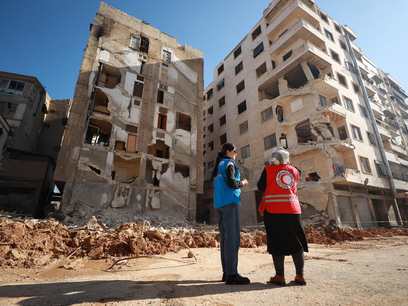 A UNHCR staff member and Red Cross staff member stand in front of a destroyed building.
