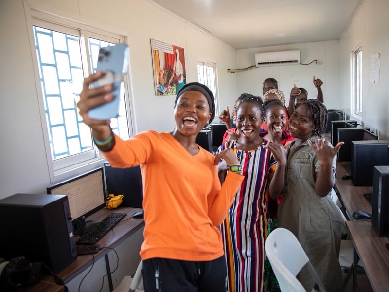 A woman in a bright orange shirt takes a selfie with a group of young women in a computer lab.