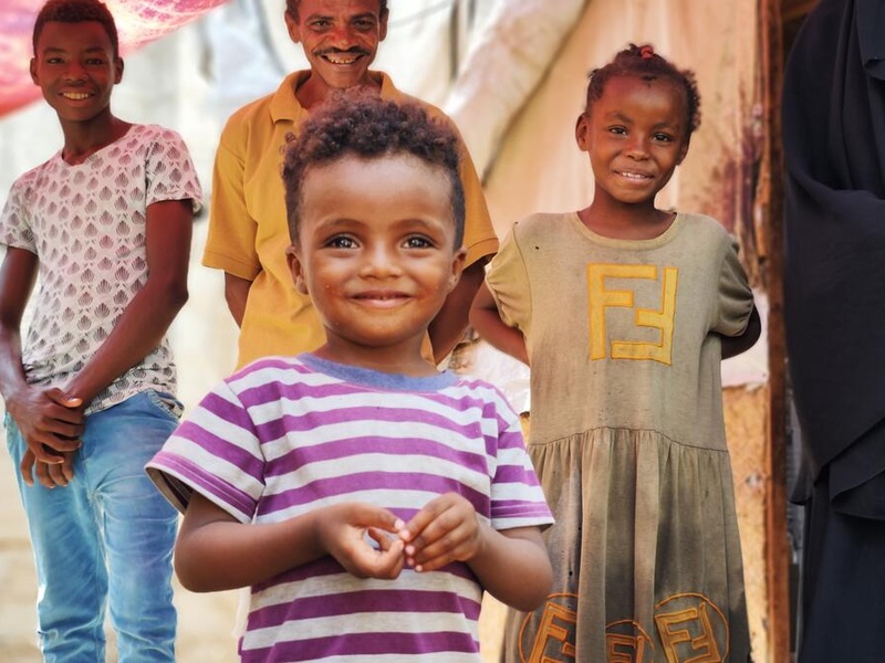 A three-year-old internally displaced boy from Yemen smiles for the camera. His father and older siblings are standing behind him.