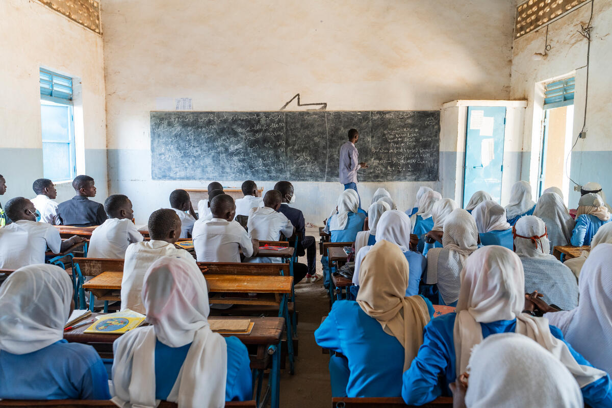 A class of Sudanese refugee students and their teacher sit in the shade of a tree.