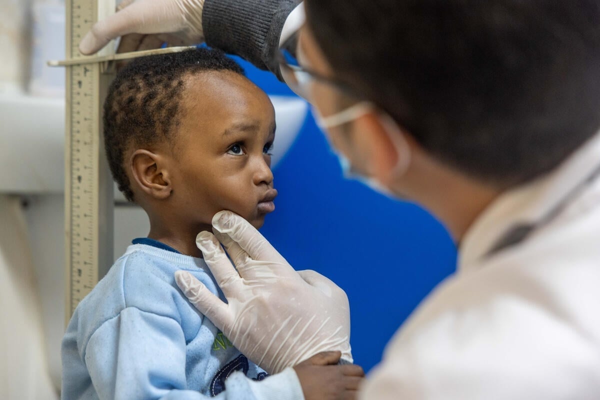 A young boy in a blue sweater stands against a height rule as a man wearing a surgical mask and gloves measures his height.