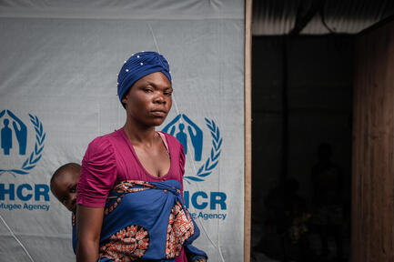 A woman carrying her baby on her back stands in front of a UNHCR shelter.