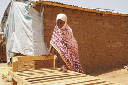 A woman constructs a wooden bed in front of a shelter made of bricks, corrugated iron and plastic sheeting.