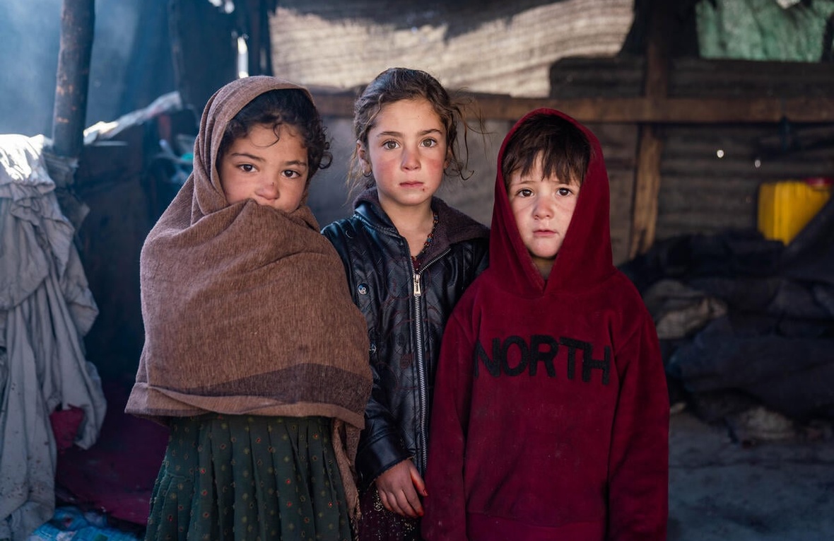 Three children stand outside their shelter in warm clothing and blankets.