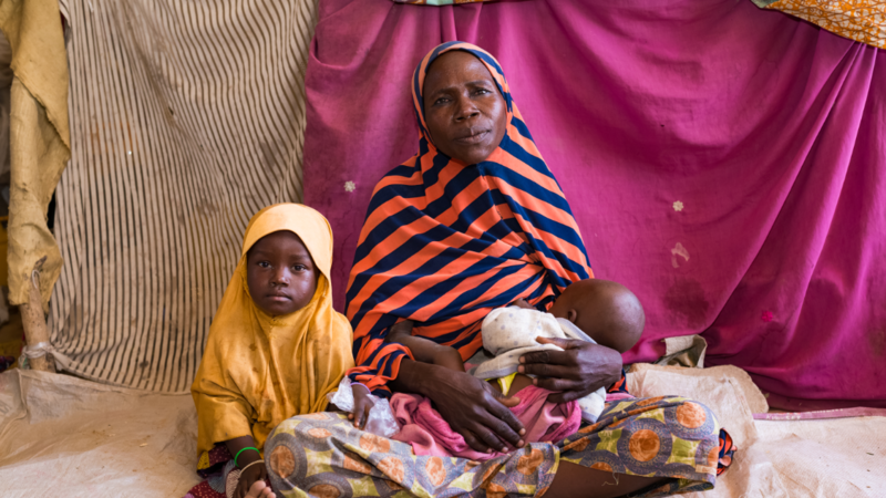 A woman holding a baby and a child both sitting on the ground.