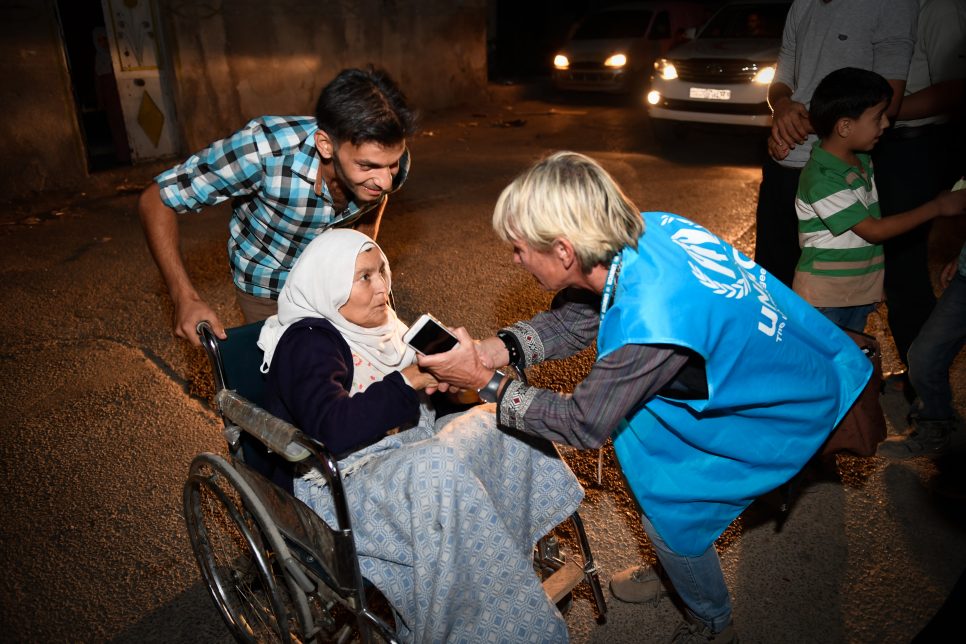 A UNHCR worker warms the hands of a woman in a wheelchair as local people come to watch aid workers offload desperately needed supplies from a convoy of inter-agency trucks in Mouadamiyeh in rural Damascus. ; After three years under siege, bombarded and starved, the rebel-held Damascus suburb of Mouadamiyeh finally surrendered to Syrian Government forces in early September 2016. A peace deal meant civilians and rebels were evacuated to other parts of the country while those who remained were able to access vital food supplies and humanitarian aid. On 22 and 24 September, a UNHCR relief convoy of trucks and staff arrived as military blockades preventing access were lifted. UNHCR staff were assisted by Syrian Arab Red Crescent volunteers and teams from other UN agencies. Despite routine blocking of UN aid, since the beginning of 2016 the UN and its partners have successfully provided assistance to 1.2 million civilians in besieged locations.