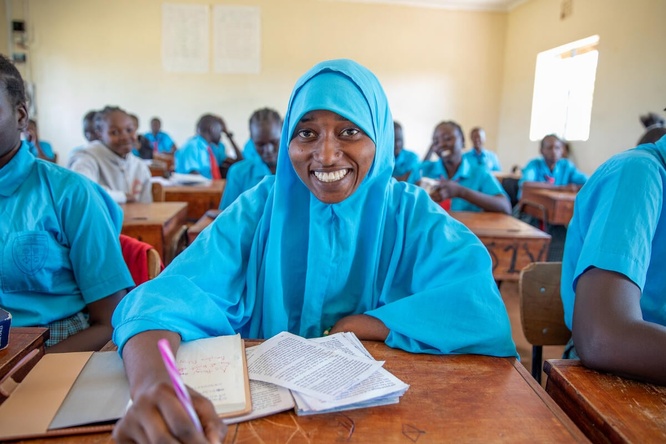A young woman wearing a headscarf sits at a desk in a classroon.