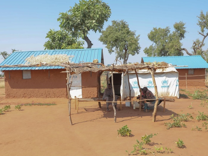 A Sudanese family sit together in the shade outside their shelter, which shows branding from UNHCR and KOICA.