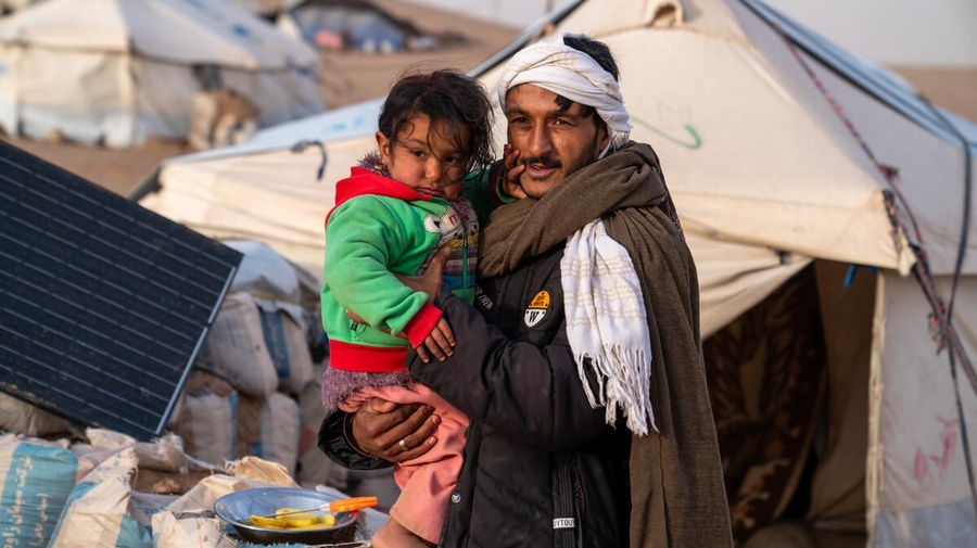 An internally displaced man in Afghanistan, wrapped in blankets, holds a small child. Behind him is a refugee tent settlement.