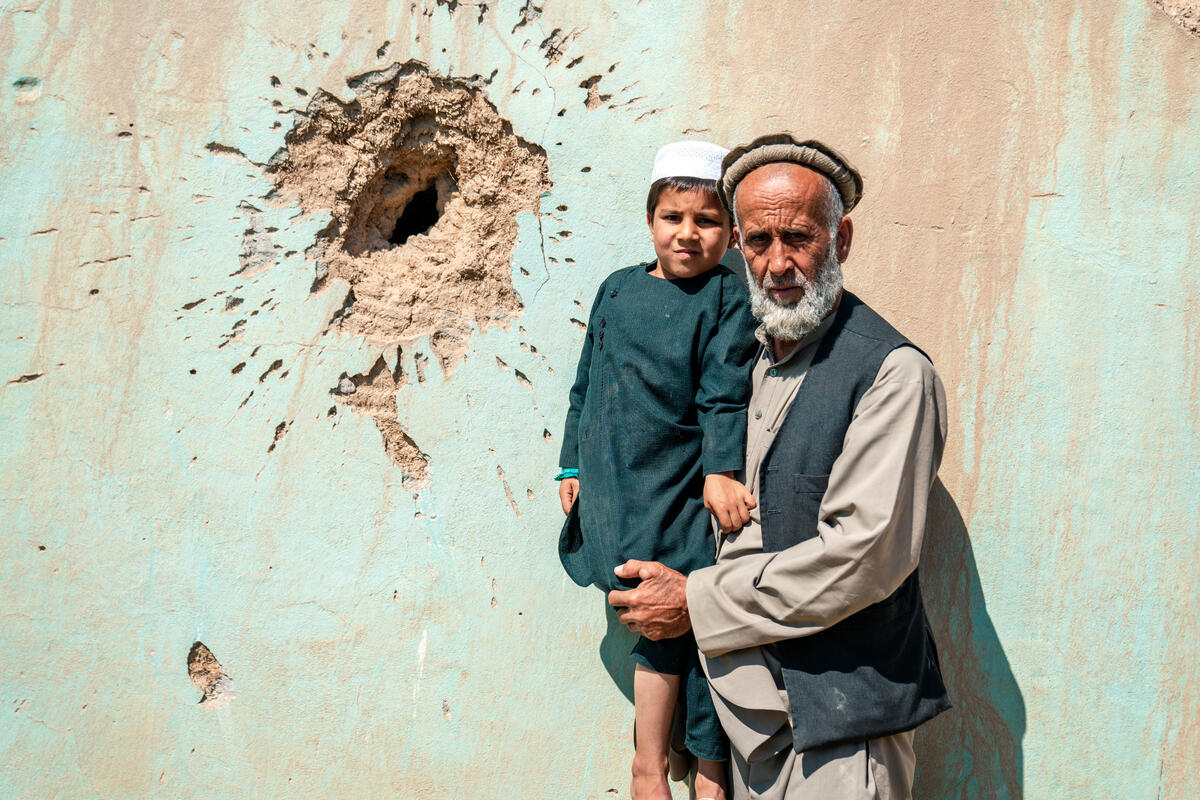 A man holds a young boy in his arms, standing in front of a wall damaged by conflict.