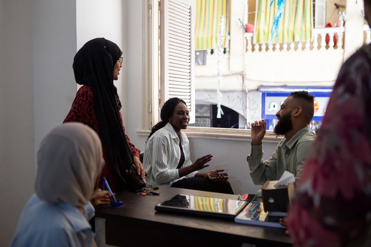 A woman and a man sit talking in front of an open window while two other women look on.