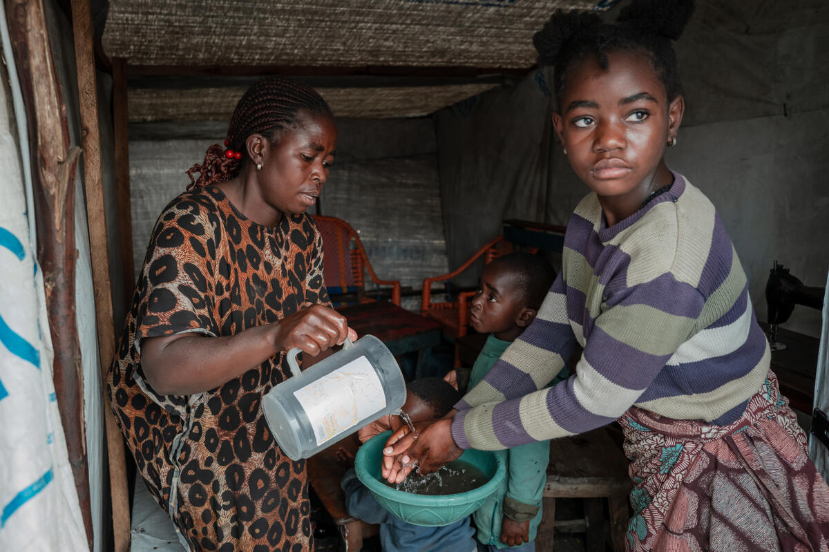 Odette helps her children wash their hands before eating a meal. A girl washes her hands in a small basin held by a woman pouring water from a plastic jug.