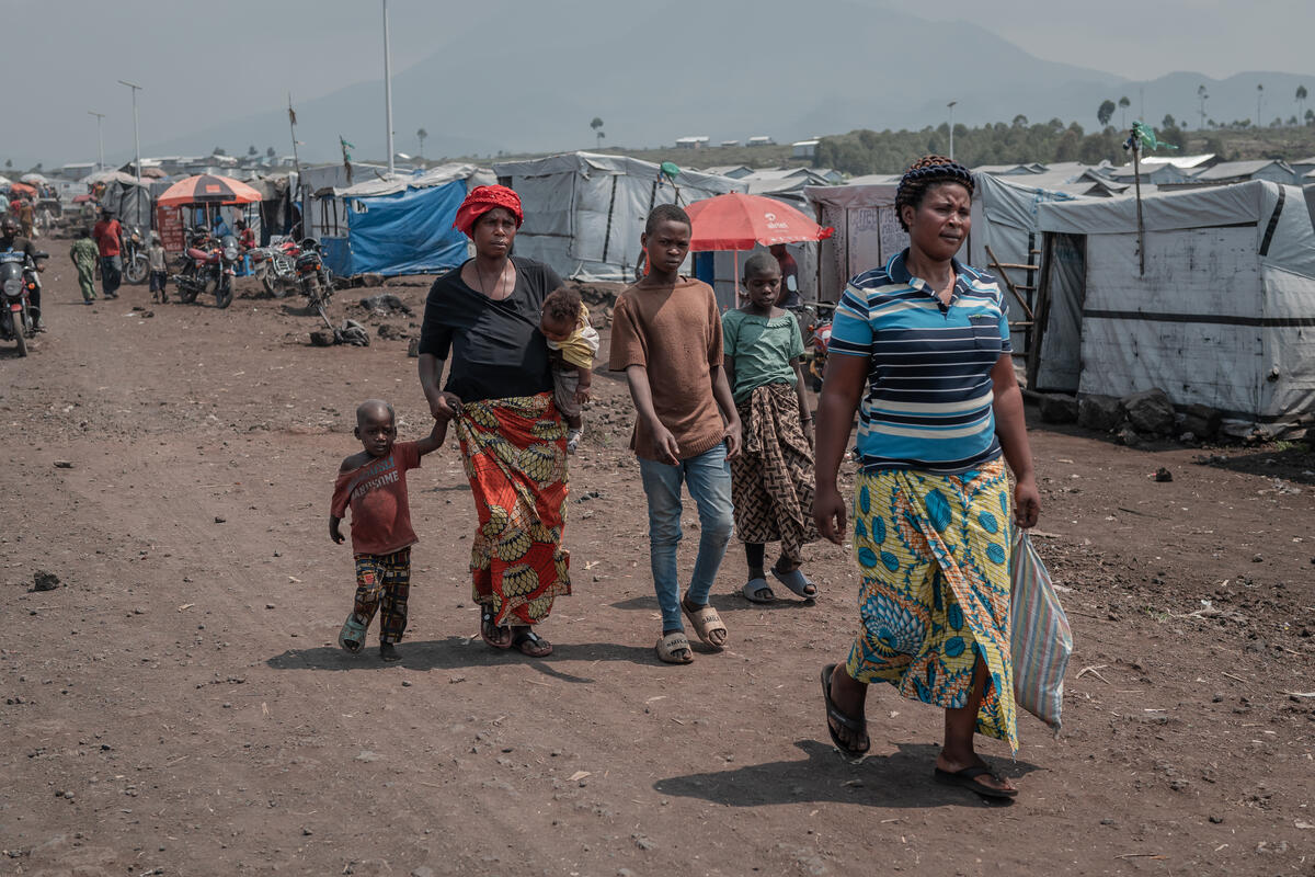 Mwamini walks through Bushagara displacement site with four of her children. A woman walks with four children down the main thoroughfare of a camp behind another woman.