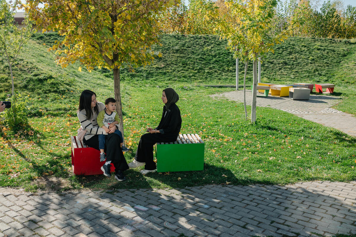 Two young women, one with a small boy on her lap, sit talking in a park.