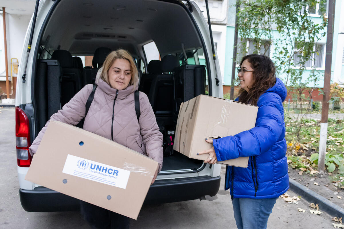 Two women wearing winter jackets unload two large boxes from a van.