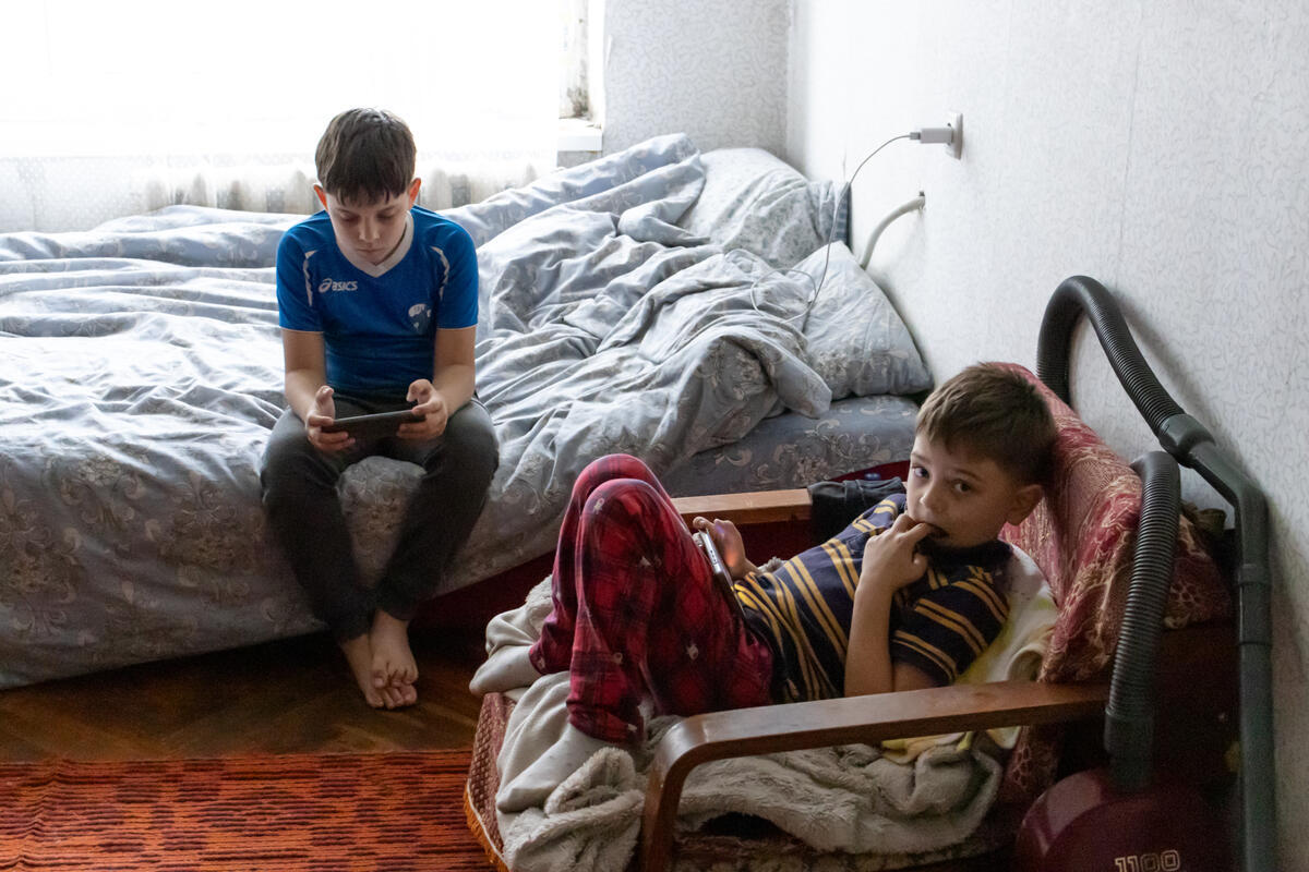 Two children play on their phones in their flat. One sits on a chair, his fingers in his mouth, looking into the camera while the other one sits on the bed.