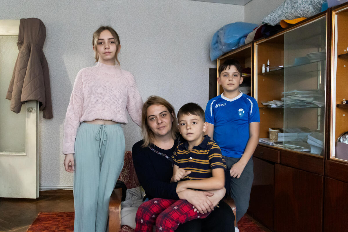 A woman poses for a photo with her three children in their house. The youngest sits on her lap while the other two stand on her sides.