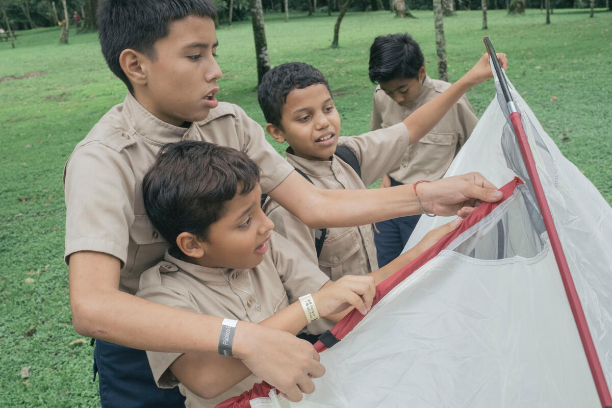 An older boy helps three younger boys wearing Scout uniforms put up a tent.