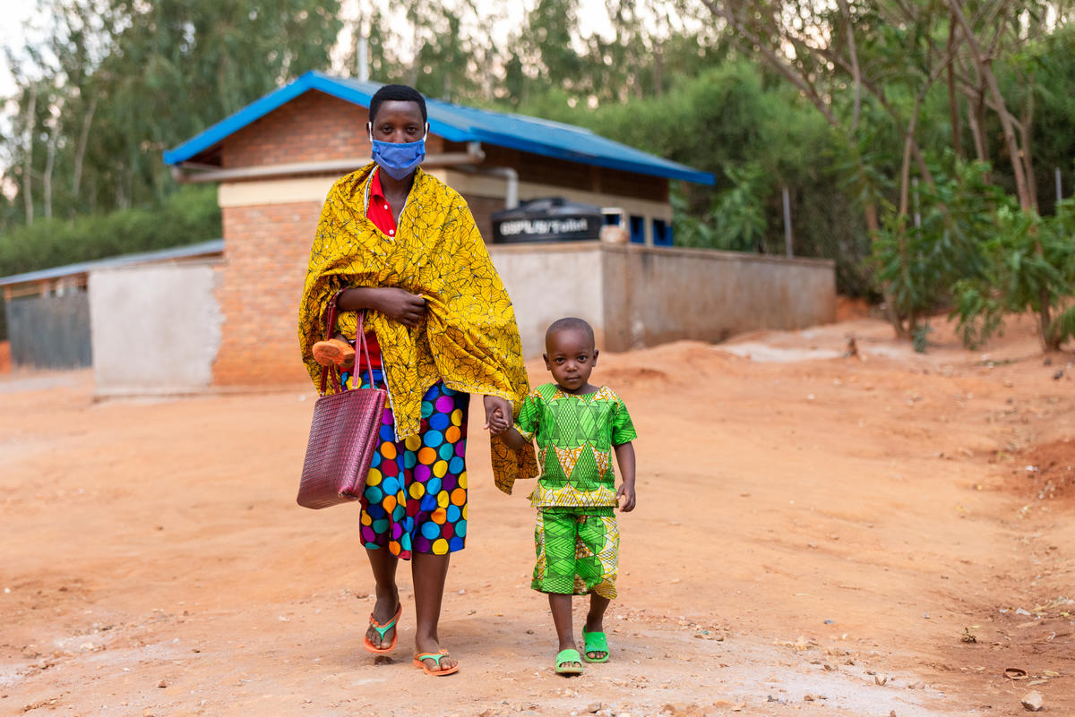 A Burundian woman wearing colourful clothes and a mask hold her child's hand.