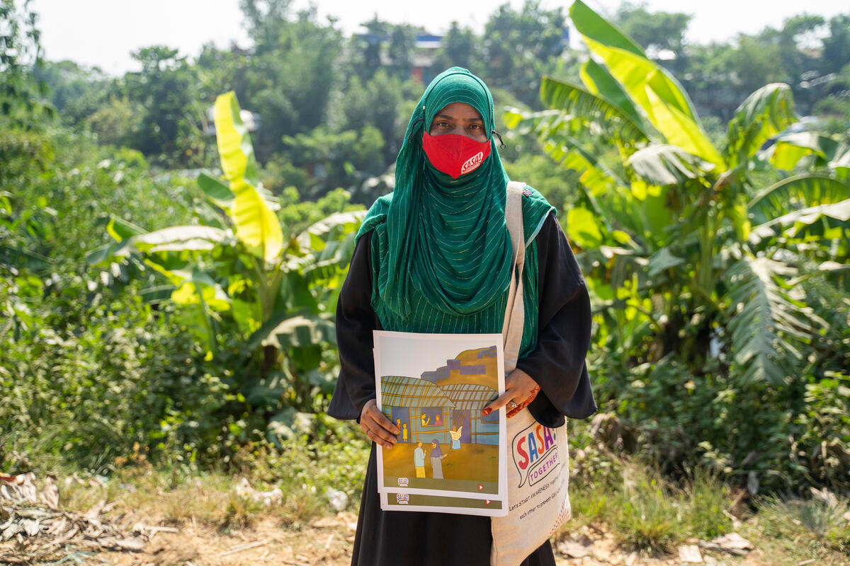 A woman wearing a hijab and holding a pair of posters looks straight into the camera. There are a number of plants behind her.