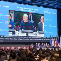 A man is pictured speaking on a giant screen in front of a conference room audience