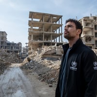 A man wearing a black UNHCR jacket looks upwards while surrounded by rubble and destroyed buidings