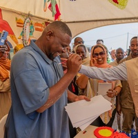 A smiling man drawing a pencil portrait on paper receives a pat on the shoulder from his smiling subject in a tent full of onlookers
