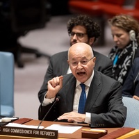 A man gestures while speaking behind a desk with people sitting in the background