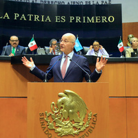 A man gestures while speaking behind a lectern with a row of people looking on from a raised platform behind