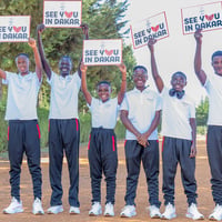Six smiling young athletes wearing training shoes and clothes hold up signs reading 'see you in Dakar' on a wide dusty road 