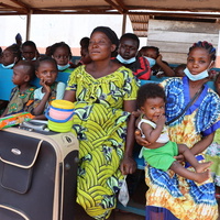 A large group of Central African refugee returnees sit on bench seating with their belongings, outside in an open-air shelter.