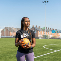 A young woman holds a football on a football pitch with rows of houses in the background