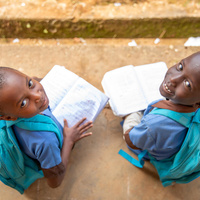 Deux jeunes écoliers en uniforme bleu et avec des sacs à dos sont assis par terre, tenant des livres ouverts tout en regardant vers le haut.