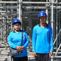 A woman and a man wearing matching blue shirts and hard hats stand outside in front of scaffolding