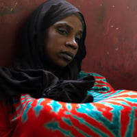 A woman in a colourful dress and a black headscarf sits on the floor leaning against a red-painted, pockmarked wall