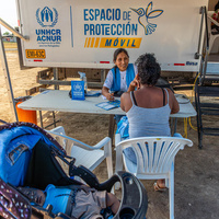 A female member of UNHCR staff speaks to a woman across a table with a baby stroller in the foreground and a truck in the background